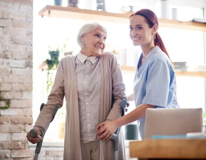 A young woman in a blue striped shirt is feeding a piece of cake to a smiling elderly woman at a table. There is a bundt cake and glasses of juice on the table.