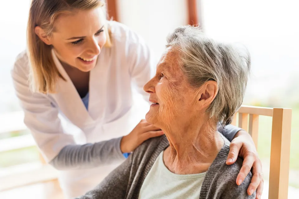 Caregiver smiling and gently supporting an elderly woman seated in a chair.